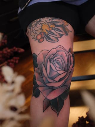 Rose tattoo with shaded petals and leaves on upper arm, with a bee design visible above on the same limb.
