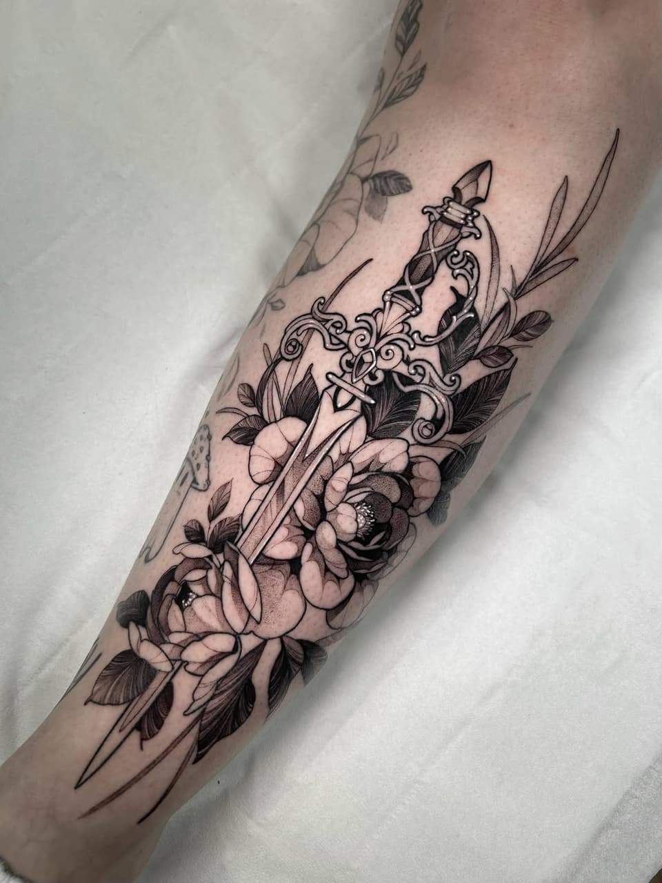 Ornate dagger with decorative scrollwork surrounded by large peonies and leafy botanicals on the forearm in black ink.
