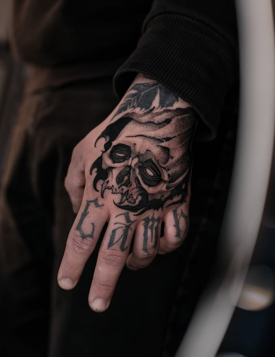 Skull tattoo with leaf details on the back of a hand, rendered in black ink with shading on tan skin.