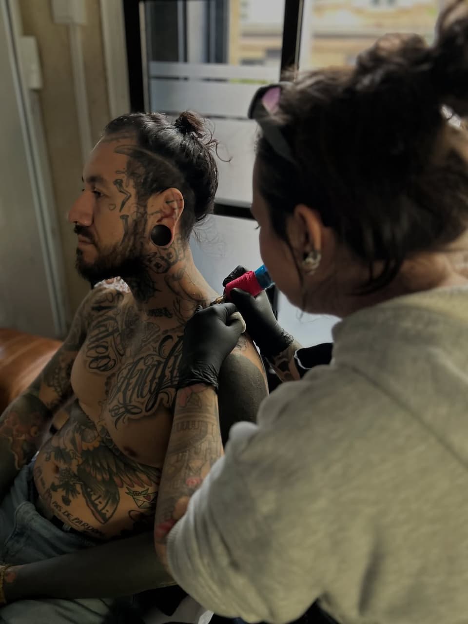 Tattoo artist applies red and black ink to client's shoulder and chest area during a studio session.