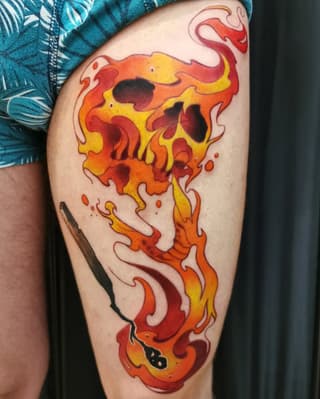 Flaming skull tattoo on thigh featuring bright red, orange, and yellow flames with black outline details and dripping elements.