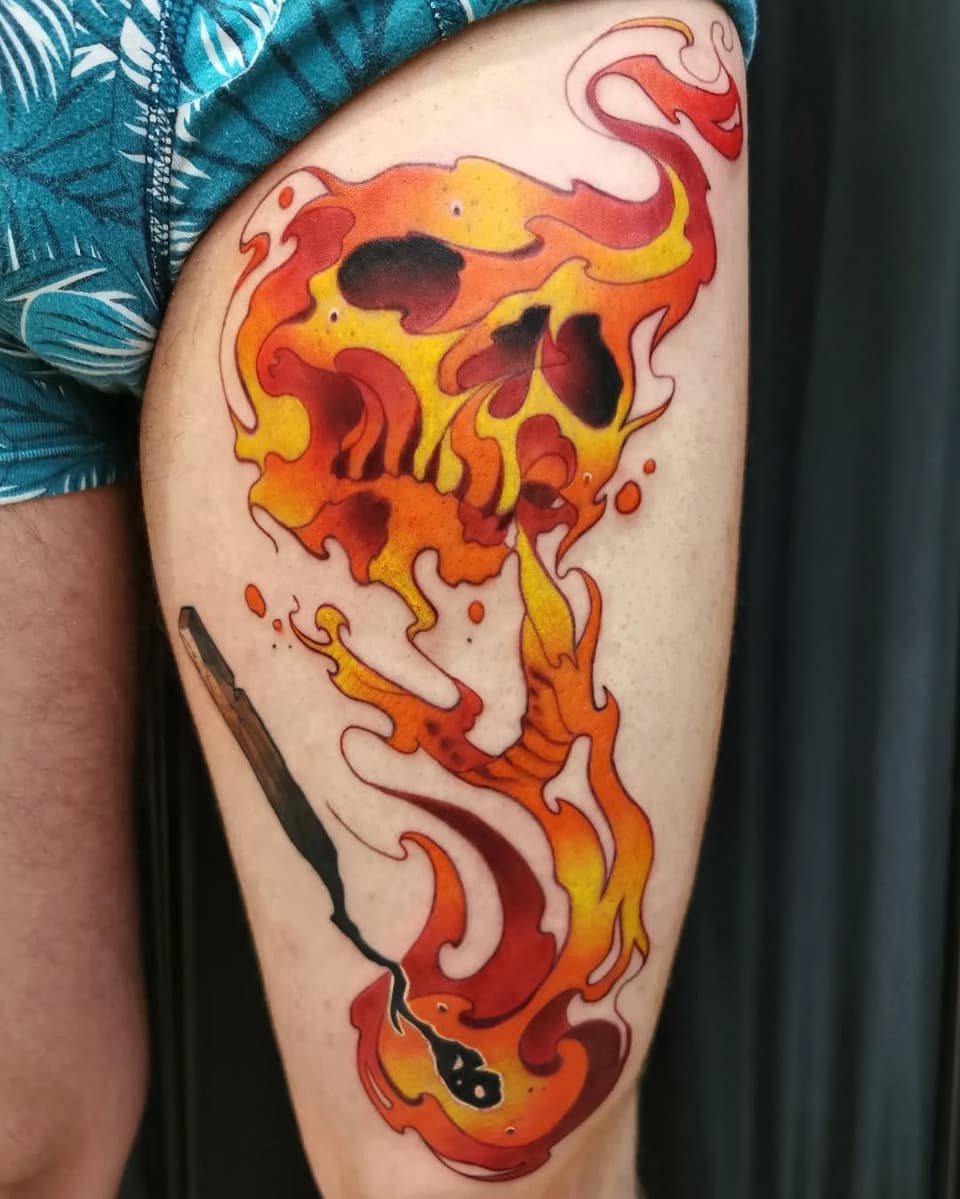 Flaming skull tattoo on thigh featuring bright red, orange, and yellow flames with black outline details and dripping elements.