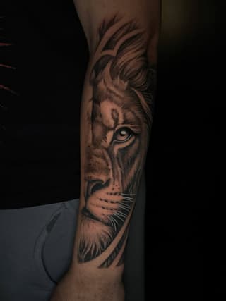 Lion head tattoo on forearm in grayscale shading with detailed facial features, flowing mane, and whiskers.