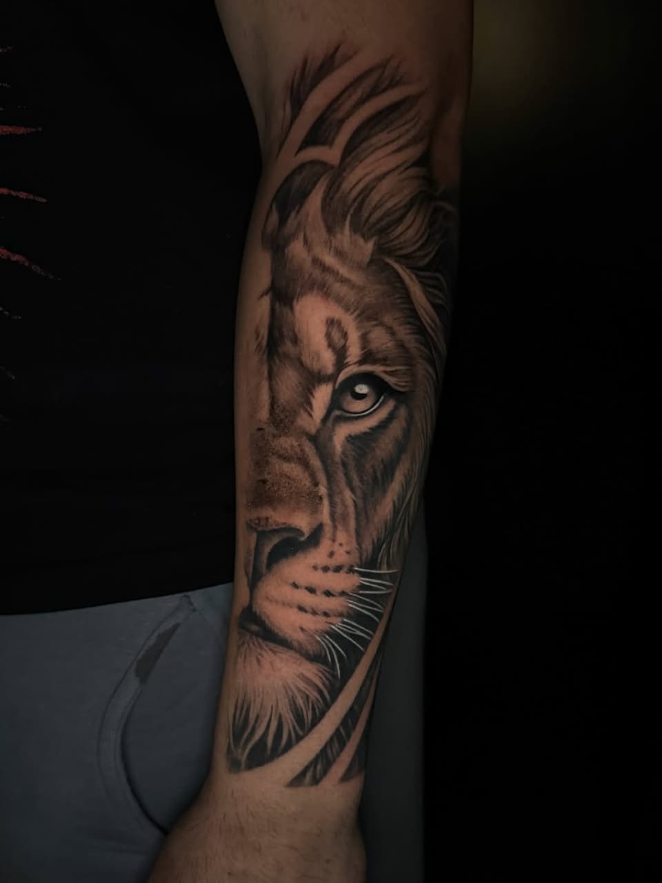 Lion head tattoo on forearm in grayscale shading with detailed facial features, flowing mane, and whiskers.