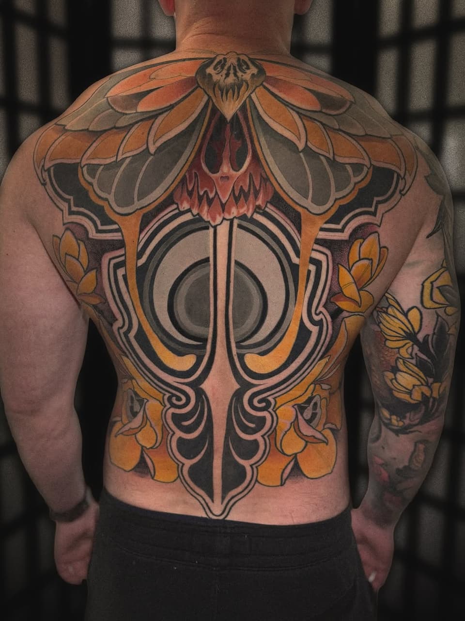 Back tattoo featuring a symmetrical design with an orange moth, central geometric circle, and ornamental flourishes in orange, black, and red ink.