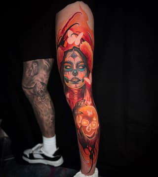 Full-leg sleeve tattoo featuring a skull-faced figure with teal eye makeup surrounded by red and orange floral elements and a glowing heart design.