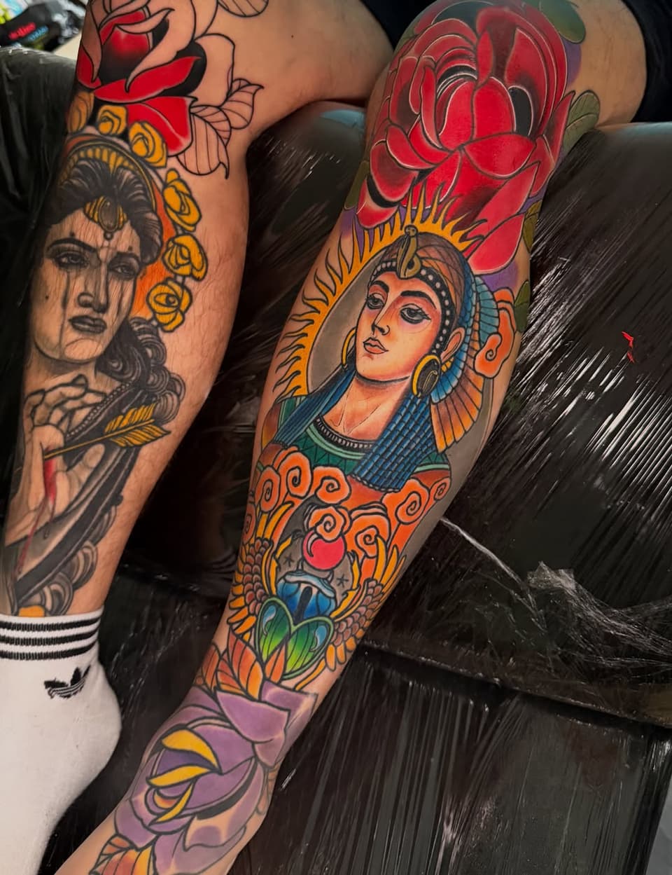 Two colorful arm tattoos featuring Egyptian-inspired portrait heads with golden headdresses, red roses, and ornate decorative elements in red, gold, blue, and purple.