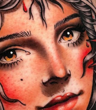 Close-up portrait tattoo of a face with golden eyes, black hair, red lips and cheeks, featuring bold black line work and warm orange-red skin tones.