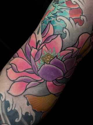 Colorful lotus flower tattoo on arm with pink, purple, and turquoise petals, detailed center, and ornamental scrollwork above and below.