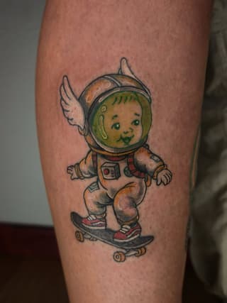 Smiling baby astronaut with wings riding a skateboard, wearing a spacesuit and helmet with green face, tattooed on a calf in tan, green, red, and black colors.
