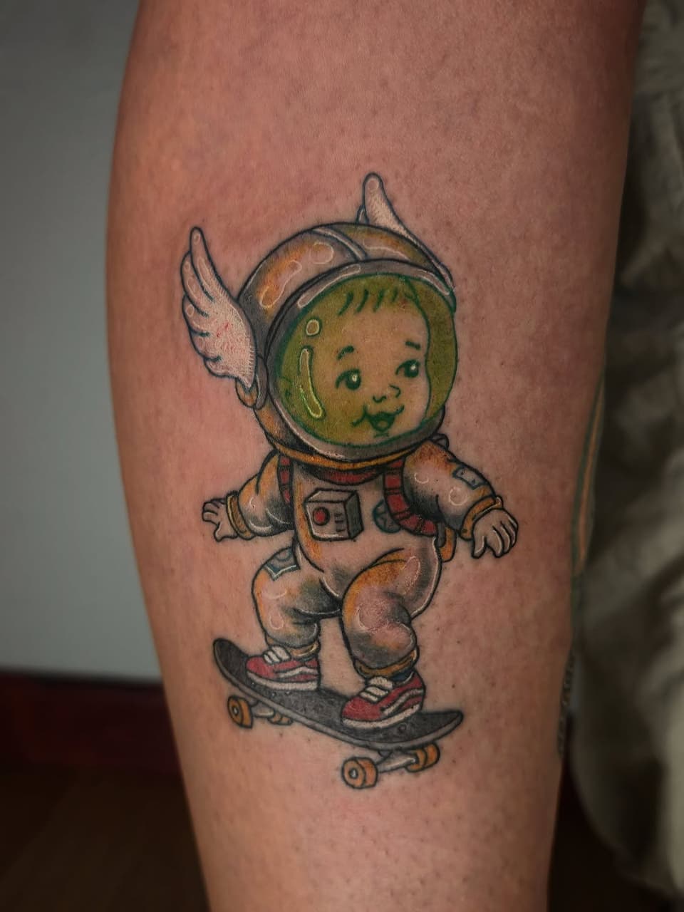 Smiling baby astronaut with wings riding a skateboard, wearing a spacesuit and helmet with green face, tattooed on a calf in tan, green, red, and black colors.