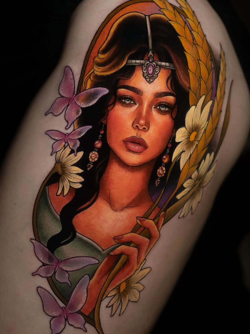 Colorful shoulder tattoo featuring a woman with a third eye jewel, surrounded by purple butterflies and yellow flowers, wearing ornate earrings.