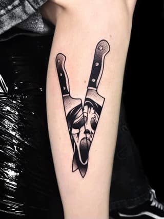 Two kitchen knives with black handles crossed through a bearded man's face in profile, tattooed on the inner forearm in black ink.