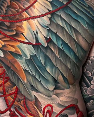 Detailed wing tattoo with layered feathers in orange, teal, and grey tones, accented with red rope designs on the arm.