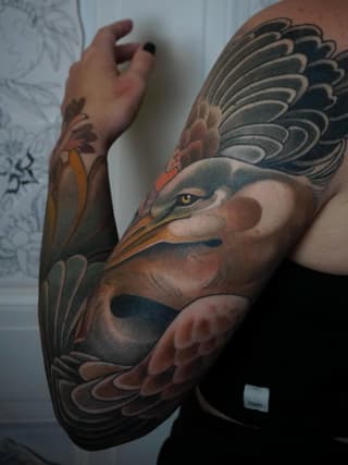 Large bird head tattoo on forearm and hand featuring detailed plumage in black, brown, orange and tan colors with a prominent eye.