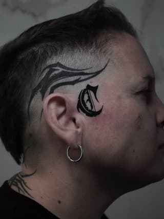 Side head tattoo featuring a curved line design above the ear and a decorative teardrop-shaped symbol with black ink below it.