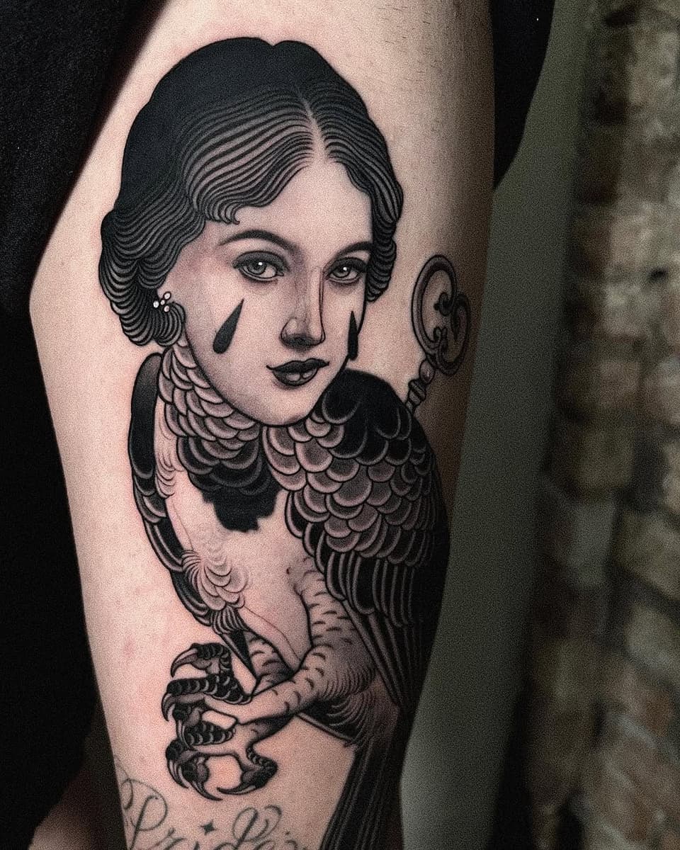 A black and white shoulder tattoo featuring a woman with wavy hair, decorative collar, and bird wings, holding taloned feet.