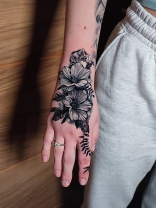 Floral tattoo on hand and forearm featuring two blooming flowers with leaves and botanical details in black ink.