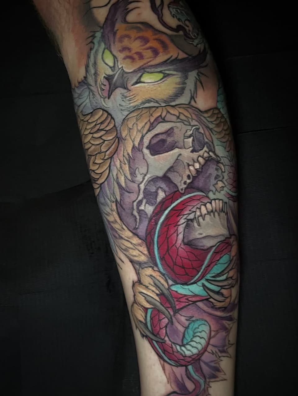 Colorful leg tattoo featuring a cat with yellow eyes at top, skull in center, and red flower with teal leaves at bottom.