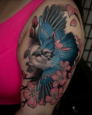 Blue jay with spread wings surrounded by pink flowers and falling petals, tattooed on the upper arm against skin.