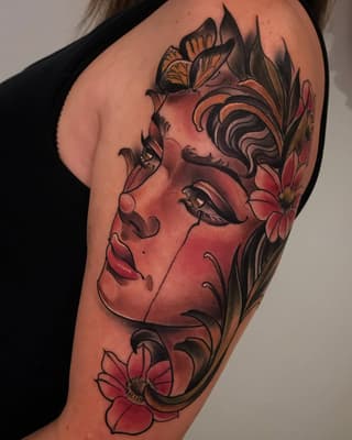 A woman's upper arm tattoo featuring a red-faced female profile surrounded by ornamental foliage, butterflies, and flowers in red and black ink.