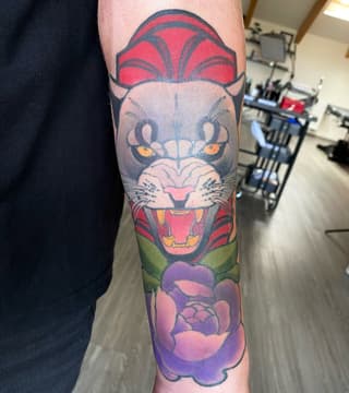 Colorful lion head tattoo on forearm featuring red mane, yellow eyes, green collar, and purple flower elements below.