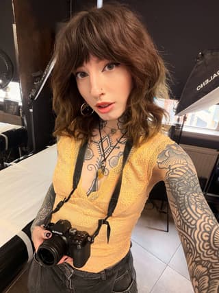 Person with brown wavy hair holding a camera, displaying detailed geometric and ornamental tattoos covering both arms and chest in black ink.