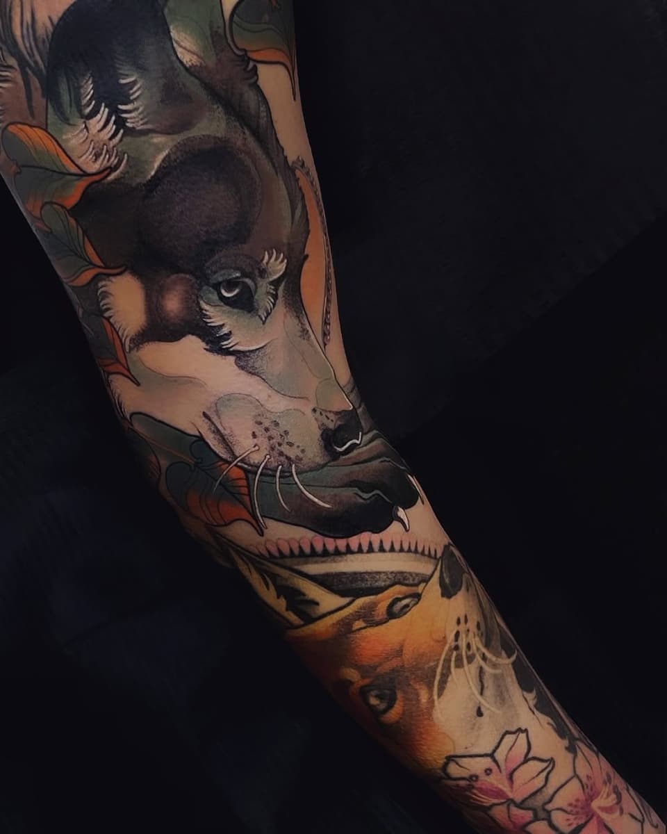 Colorful arm tattoo featuring stacked animal faces including a deer, fox, and fish with botanical elements in orange, black, and natural tones.