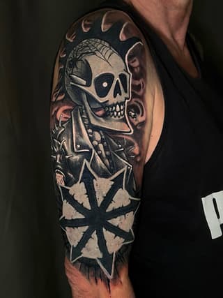 Skull with skeletal hand and geometric cross design tattooed on upper arm in black and grey tones with shading.