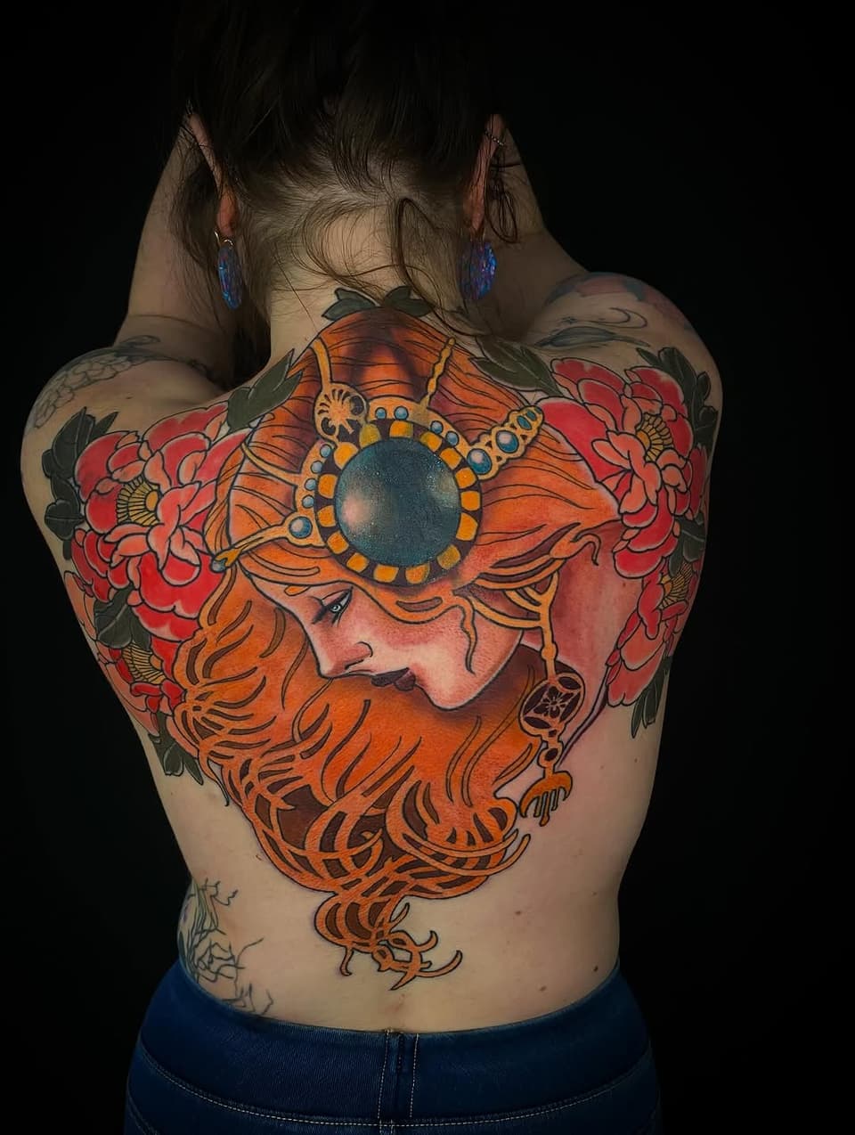Back tattoo featuring a bearded man's profile with orange hair and beard, surrounded by red flowers and a large blue circular jewel ornament.