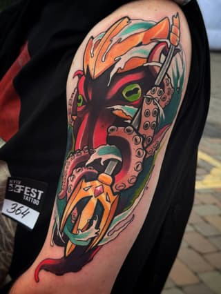 Colorful upper arm tattoo featuring a demon face with green eyes, orange and red coloring, tentacle-like appendages, and ornamental details.