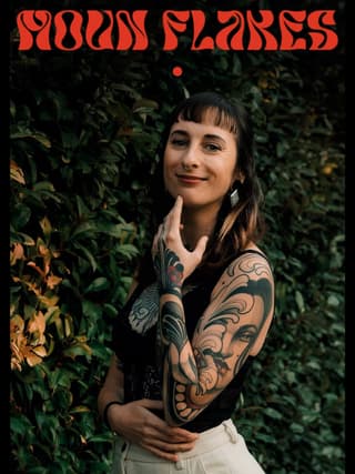 Tattooed woman with elaborate sleeve designs featuring ornamental scrollwork and a face portrait on her arm, posed against green foliage.