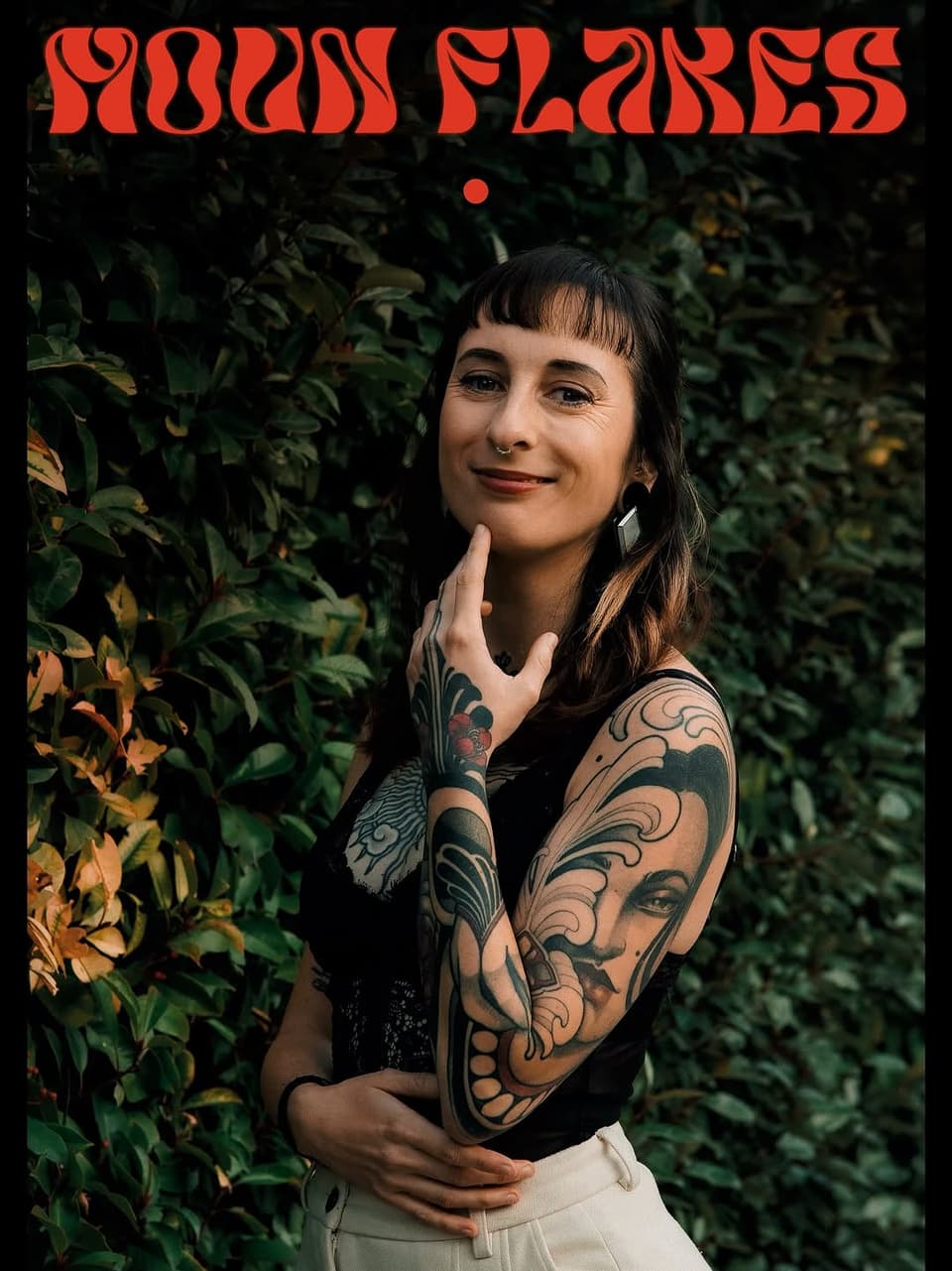Tattooed woman with elaborate sleeve designs featuring ornamental scrollwork and a face portrait on her arm, posed against green foliage.