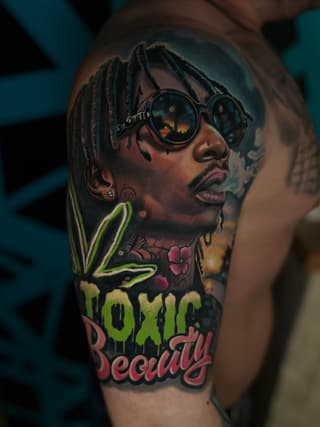Colorful shoulder tattoo featuring a man with dreadlocks wearing round sunglasses, cannabis leaf, and "Toxic Beauty" text in green and red.