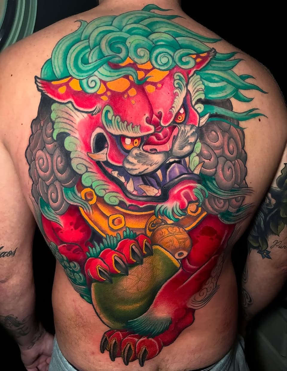 Colorful lion head tattoo on upper back featuring red, green, orange, and gold tones with ornamental details and a pendant held in its paws.