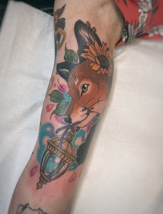 Colorful deer head tattoo with sunflower, lantern, and botanical elements on the forearm in orange, pink, teal, and green tones.