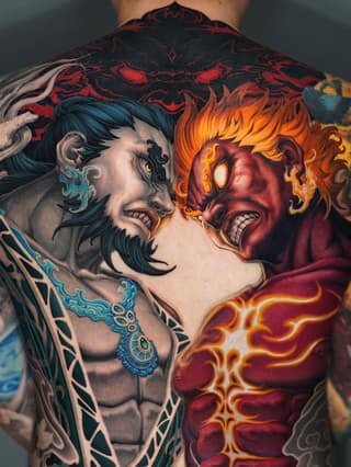 Back tattoo featuring two demonic faces in blue and red/orange tones with ornate decorative patterns and geometric designs across the shoulders and torso.
