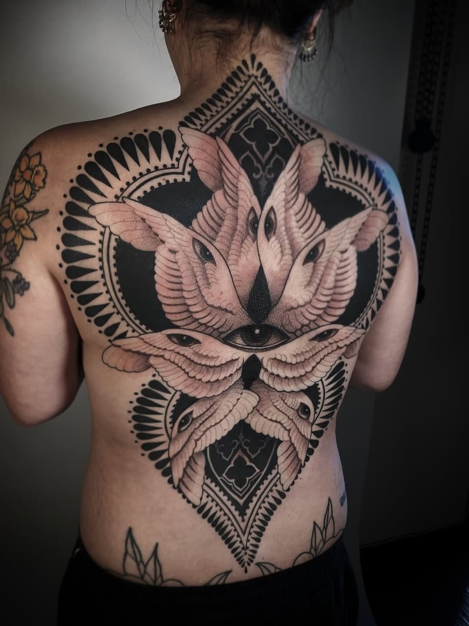 Back tattoo featuring three winged birds surrounding geometric diamond shapes with decorative fan patterns in black and pink ink.