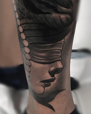 Close-up side profile portrait tattoo on forearm showing a face with closed eyes, lips, and decorative circular elements along the upper shoulder area in black and grey ink.