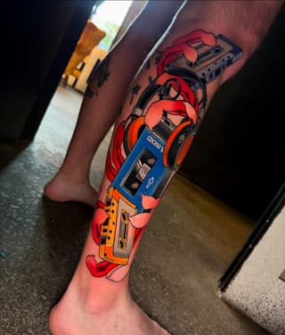 Colorful stacked cassette tape tattoo on a leg featuring red, blue, and yellow Sony and other branded tapes with decorative red ribbon elements.