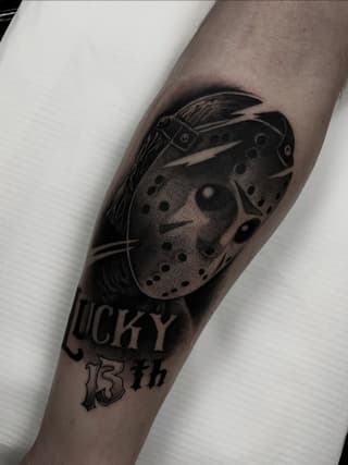 Hockey mask with buckle strap and holes tattooed on forearm with "LUCKY" and "13th" text below in black ink.