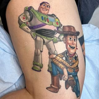 Colorful tattoo on upper arm featuring two animated characters: a space ranger in green and white suit holding a cowboy in brown plaid shirt and hat.