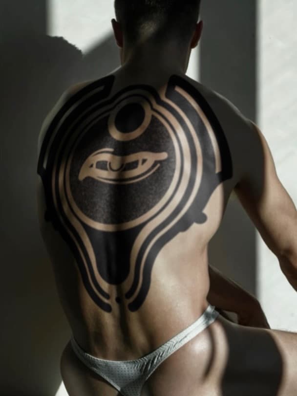 Shirtless man photographed from behind displaying a large geometric tattoo featuring concentric circles and curved lines in black and tan ink across his upper back.