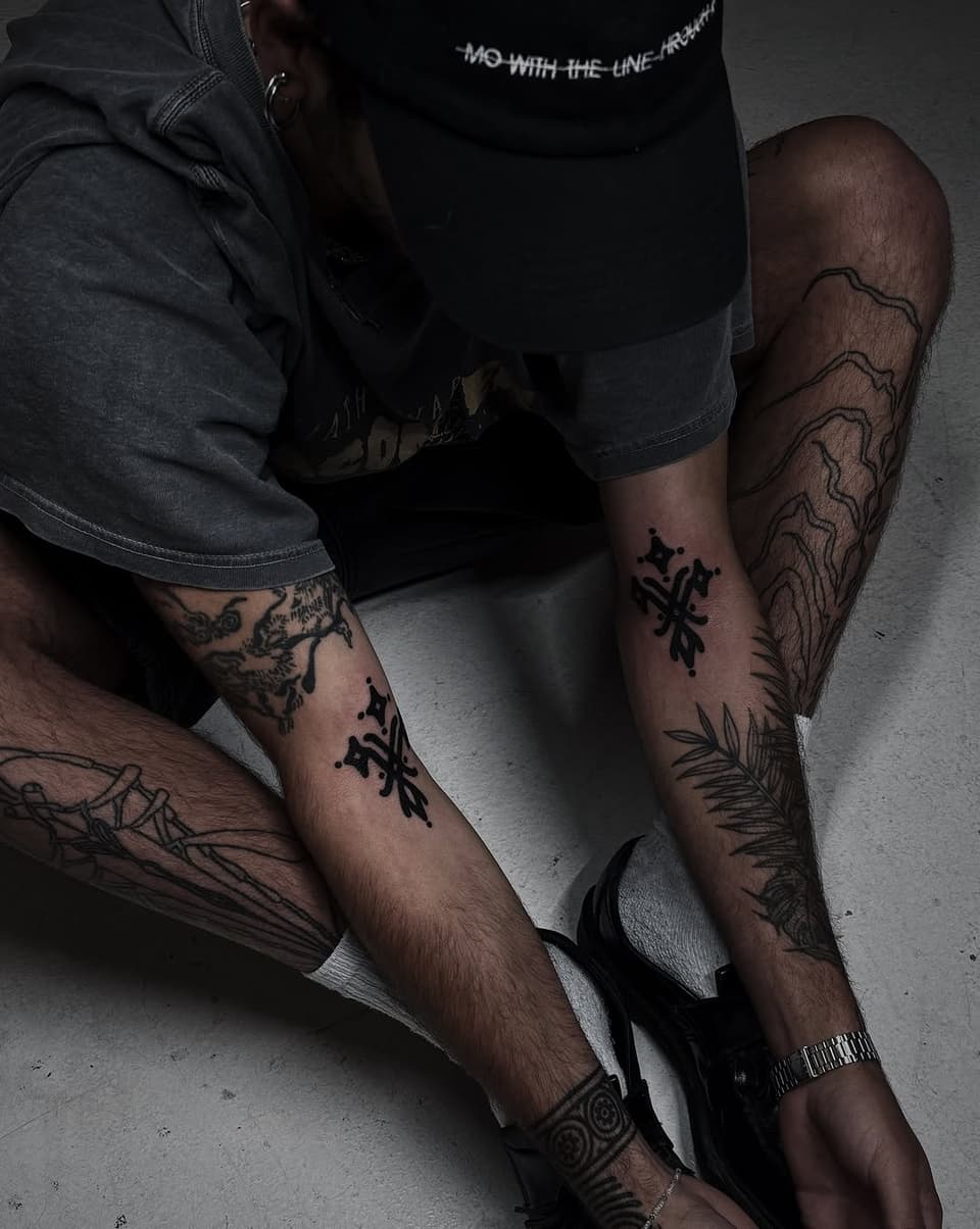 Person displaying multiple black ink tattoos on both arms and legs, including geometric patterns, botanical elements, and symbolic designs on light skin tone.