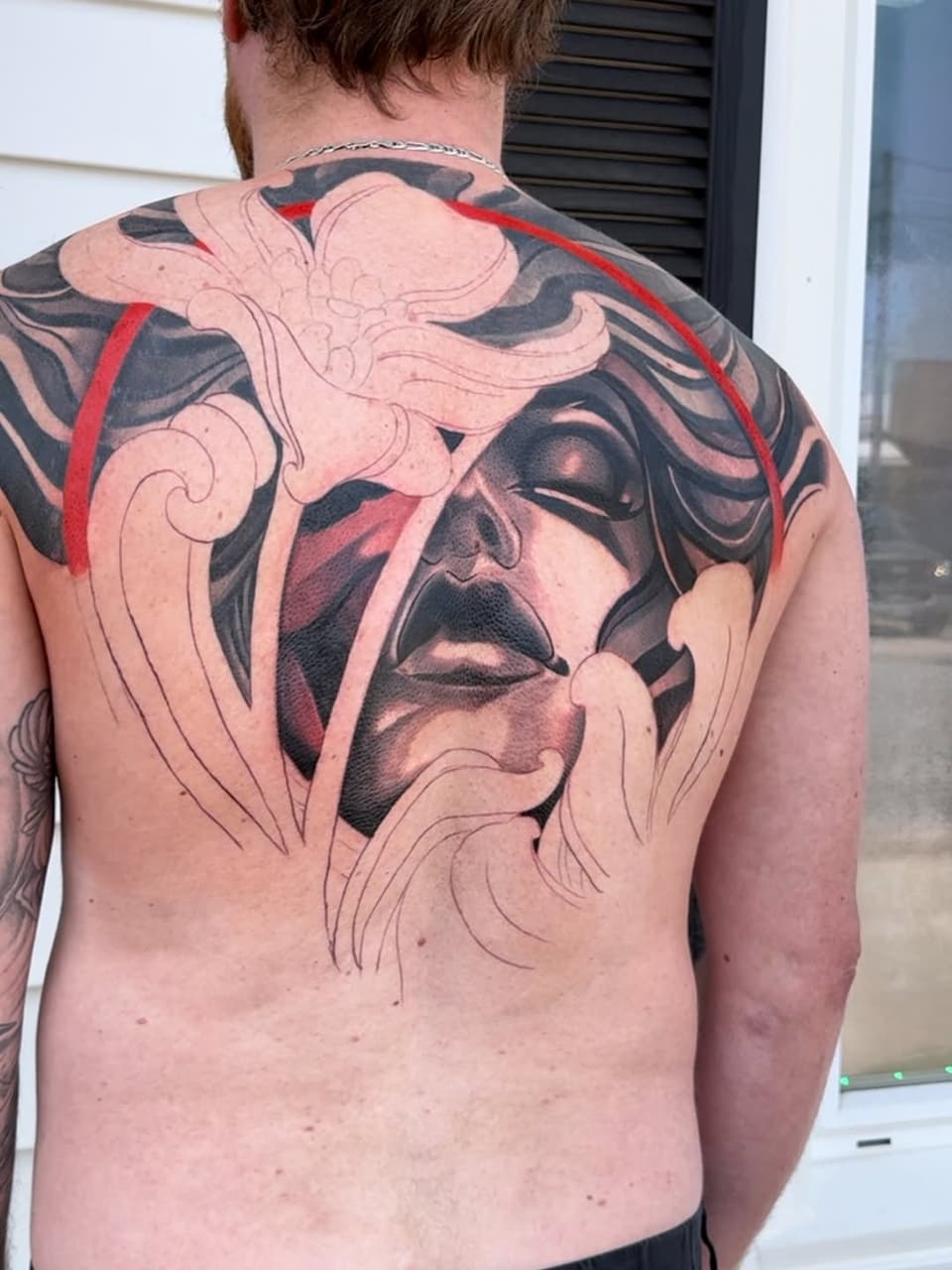 Large-scale portrait tattoo covering the back featuring a face in black and red ink with ornamental swirling designs in skin tone surrounding it.
