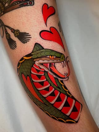 Colorful snake tattoo on forearm featuring a green head with orange and red striped body, red hearts above, and birds in upper background.