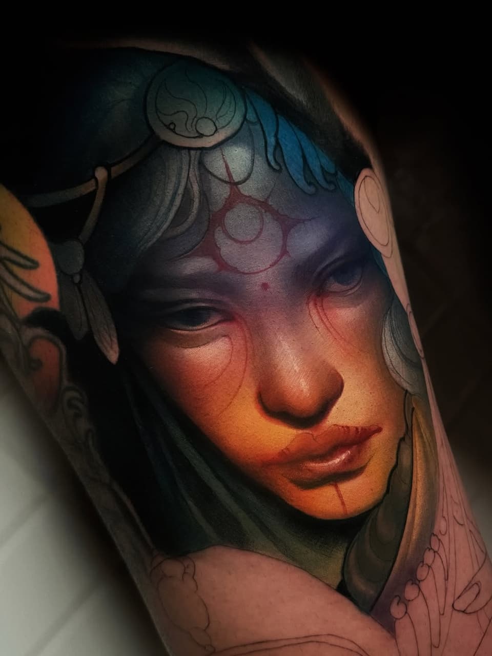 Colorful portrait tattoo on upper arm featuring a woman's face with blue and red facial markings, ornate headdress, and decorative line work.