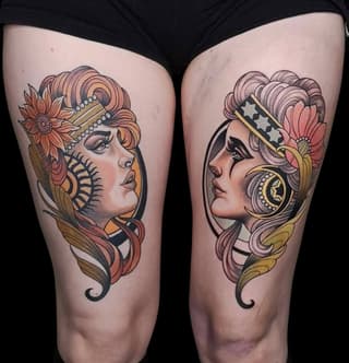 Two complementary thigh tattoos of stylized female heads with ornate headdresses, flowing hair, and decorative elements in coral, gold, and black ink.