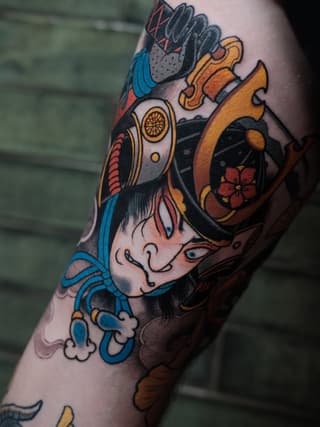 Colorful tattoo on an arm featuring a stylized face with blue and gold ornamental elements, holding a decorative object with red flowers and gold accents.