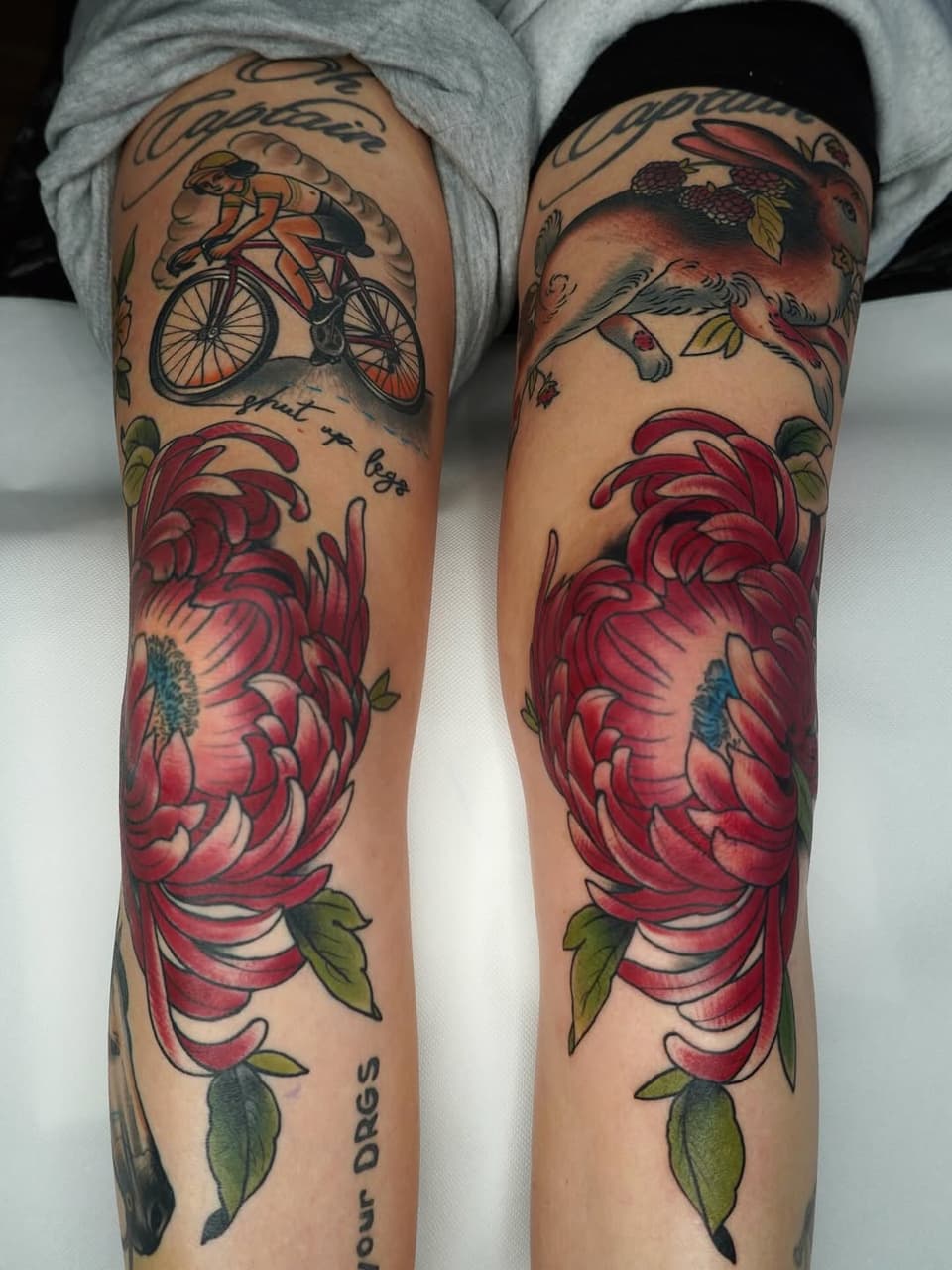 Two legs displaying matching floral leg sleeve tattoos featuring large red peonies, bicycles, small animals, and green leaves in color.